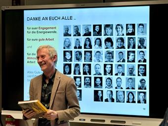 Ein Mann im braunen Sakko steht lächelnd vor einem großen, digitalen Whiteboard, das eine Präsentationsfolie zeigt. Er hält ein offenes Buch in den Händen. Die Folie zeigt den Text "DANKE AN EUCH ALLE...". Darunter befindet sich eine Collage aus vielen Schwarz-Weiß-Porträtfotos des Teams, angeordnet in einem Raster.