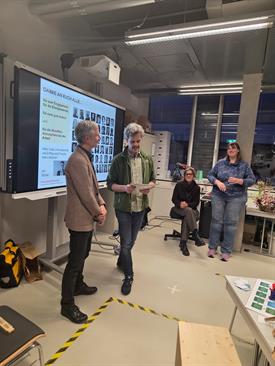 Eine Weitwinkel-Aufnahme einer Präsentation in einem modernen Seminarraum. Links steht derselbe ältere Mann im braunen Sakko vor einem Whiteboard und hört zu. Mittig steht ein jüngerer Mann mit grauen, lockigen Haaren und grünem Oberteil, der einen Text von einem Blatt Papier abliest. Im Hintergrund sitzen zwei Frauen auf Bürostühlen: eine links in Schwarz, lächelnd, und eine rechts in einem dunkelblauen, gemusterten Oberteil, die lachend ein Glas hält. Im Vordergrund sind Tische mit Arbeitsmaterialien und eine gelb-schwarze Markierung am Boden zu sehen. Die Decke zeigt freiliegende Rohre und moderne Beleuchtung.