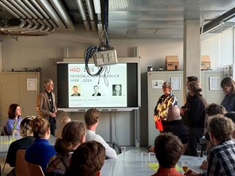 Eine Perspektive von einem Publikumstisch aus auf eine formelle Veranstaltung. Im Hintergrund, vor einem Whiteboard, steht ein älterer Mann im braunen Sakko und lächelt. Links von ihm, hinter einem separaten Tisch, sitzt eine Frau in violetter Kleidung. Rechts vom Mann steht eine Frau in einer schwarz-weiß gemusterten Bluse und spricht. Um sie herum stehen mehrere Personen. Das Whiteboard zeigt eine Folie mit dem HSD ZIES-Logo und dem Titel "PERSÖNLICHER RÜCKBLICK 1998 ... 2026", darunter drei Porträtfotos desselben Mannes in verschiedenen Lebensabschnitten mit Jahreszahlen. Das Datum "Abschiedsfeier, 26. Februar 2026" ist zu lesen. Im Vordergrund sind die Hinterköpfe und Schultern der sitzenden Zuhörer an Tischen zu sehen. Die Decke ist voller komplexer Industrie-Rohrleitungen.