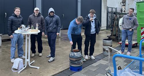 Mehrere Studierende stehen im Freien neben einem Brennholzkocher aus einem umgebauten Metallfass und einem Stehtisch mit Grillzutaten und Getränken auf dem Hof einer Hochschule.