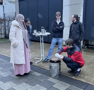 Studierende stehen draußen in Winterkleidung um einen improvisierten Brennholzkocher und einen Stehtisch mit Thermoskannen und Snacks auf einem Hochschulgelände.