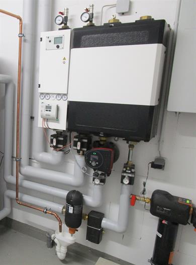 Technische Heizungs- und Sanitäranlage mit Pumpen, Druckmessern, Rohrleitungen und Steuerungseinheiten an einer Wand.
