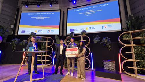 Weitwinkelaufnahme der Konferenzbühne. Zwei große Bildschirme zeigen die Gewinner des „SEED BEST EXPERT ARTICLE AWARD 2026“: Julian Spratte, Santatrinilaina Josiah Andriamanahazoarilala und Franziska Schaube. Die Preisträger stehen mit der Auszeichnung auf der Bühne.