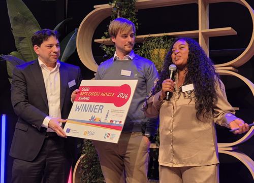 Fröhliche Szene auf der Bühne: Ein Mann im dunklen Anzug übergibt die große „Winner“-Tafel an einen Kollegen im blauen Pullover, während eine Frau daneben herzlich lacht. Im Hintergrund ist moderne Dekoration aus Holzregalen und Pflanzen zu sehen.