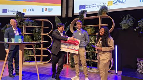 Drei Personen stehen auf einer Bühne und präsentieren eine große Urkunde mit der Aufschrift „SEED BEST EXPERT ARTICLE AWARD 2026 WINNER“. Eine Frau mit langen, lockigen Haaren spricht dabei in ein Mikrofon.