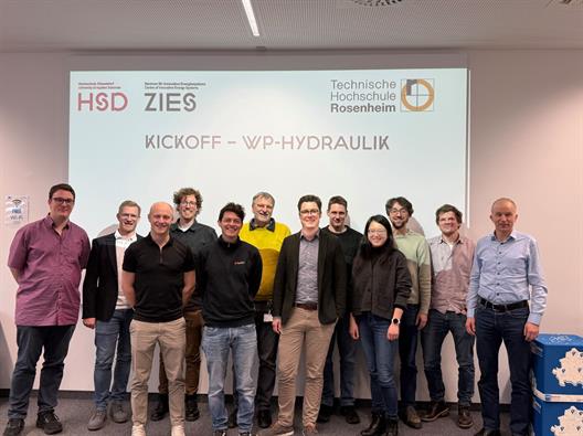 Mehrere Personen stehen dicht nebeneinander in einem Seminarraum vor einer hellen Projektionswand und blicken zur Kamera; auf der Leinwand sind die Logos der Hochschule Düsseldorf, des Zentrums für Innovative Energiesysteme und der Technischen Hochschule Rosenheim sowie der Titel „Kickoff – WP-Hydraulik“ zu sehen.