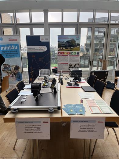 Ein Infostand der Hochschule Düsseldorf, der sich auf innovative Energiesysteme konzentriert. Der Stand ist mit verschiedenen Plakaten, Broschüren und technischen Geräten ausgestattet. Ein Schild auf dem Tisch informiert über das 'Zdi-Schülerinnenlabor – Energiewende macht Schule'. Der Stand befindet sich in einem hellen, modernen Raum mit großen Fenstern.